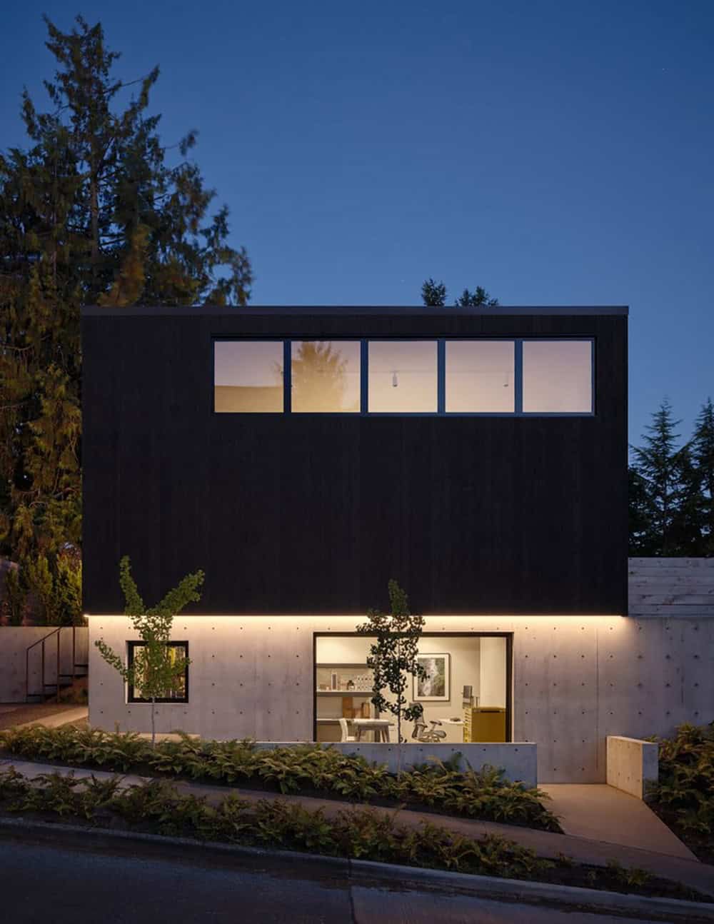 Night exterior of two-story dark wood and concrete home with clerestory windows and glowing lower-level studio