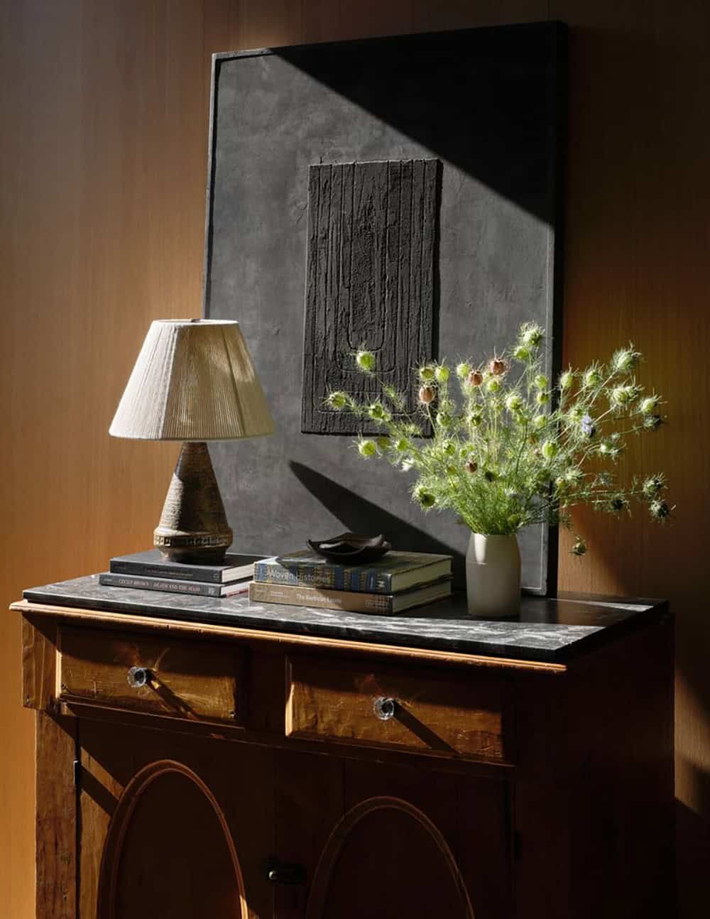Antique pine sideboard with black marble top, textured dark artwork, table lamp, and thistle arrangement