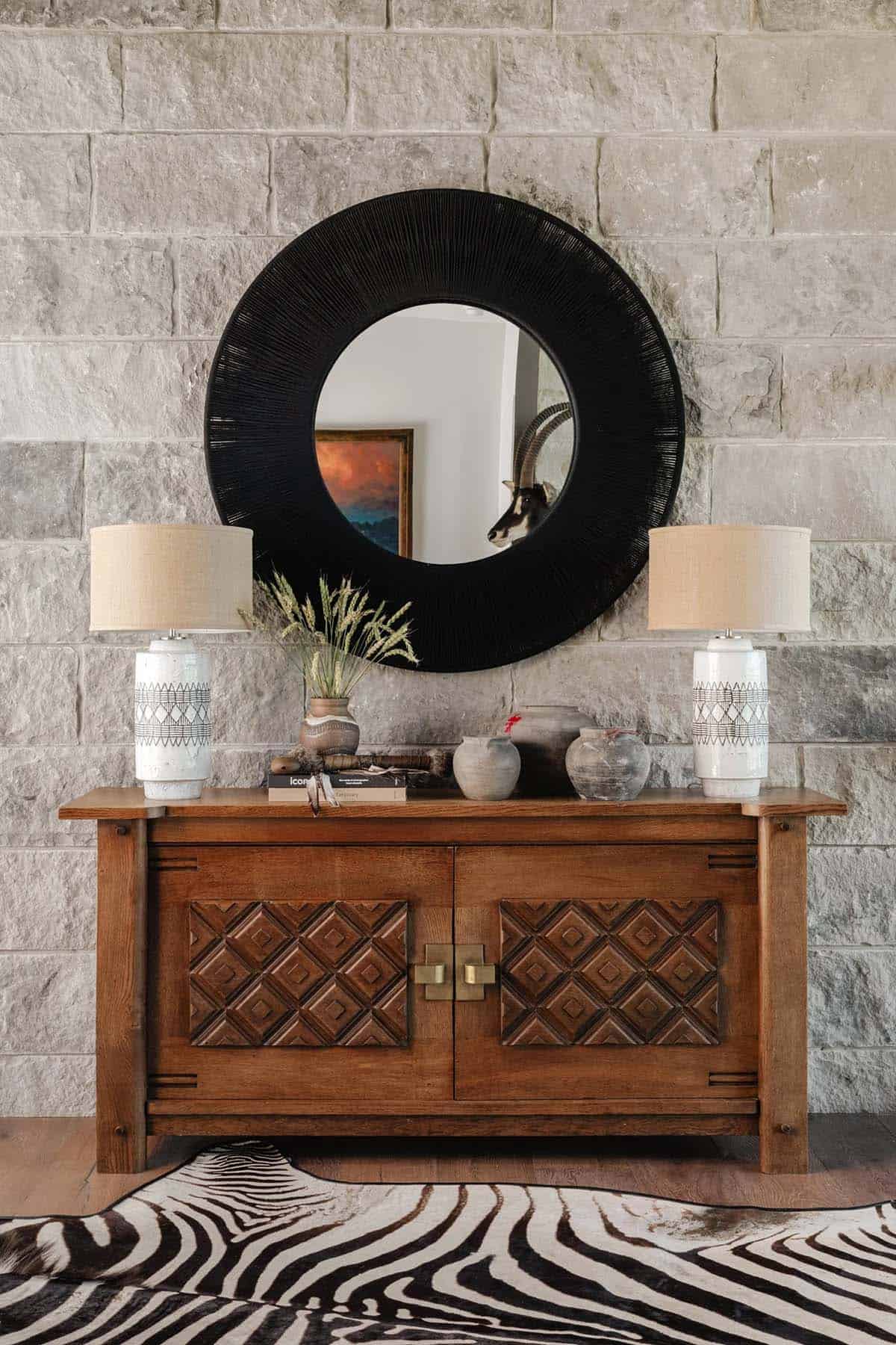 Rustic wood console with decorative lamps and round black mirror against limestone wall