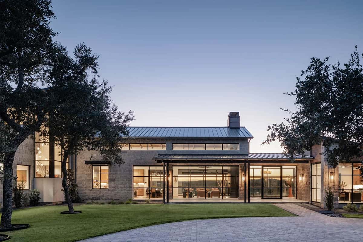 Hill Country ranch home illuminated at dusk with steel frame windows and mature oak trees