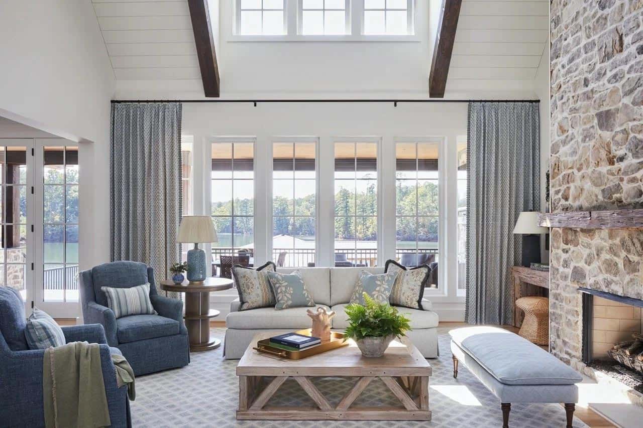 Bright lake house living room with vaulted ceiling, exposed wood beams, stone fireplace, blue armchairs, and large windows overlooking the water