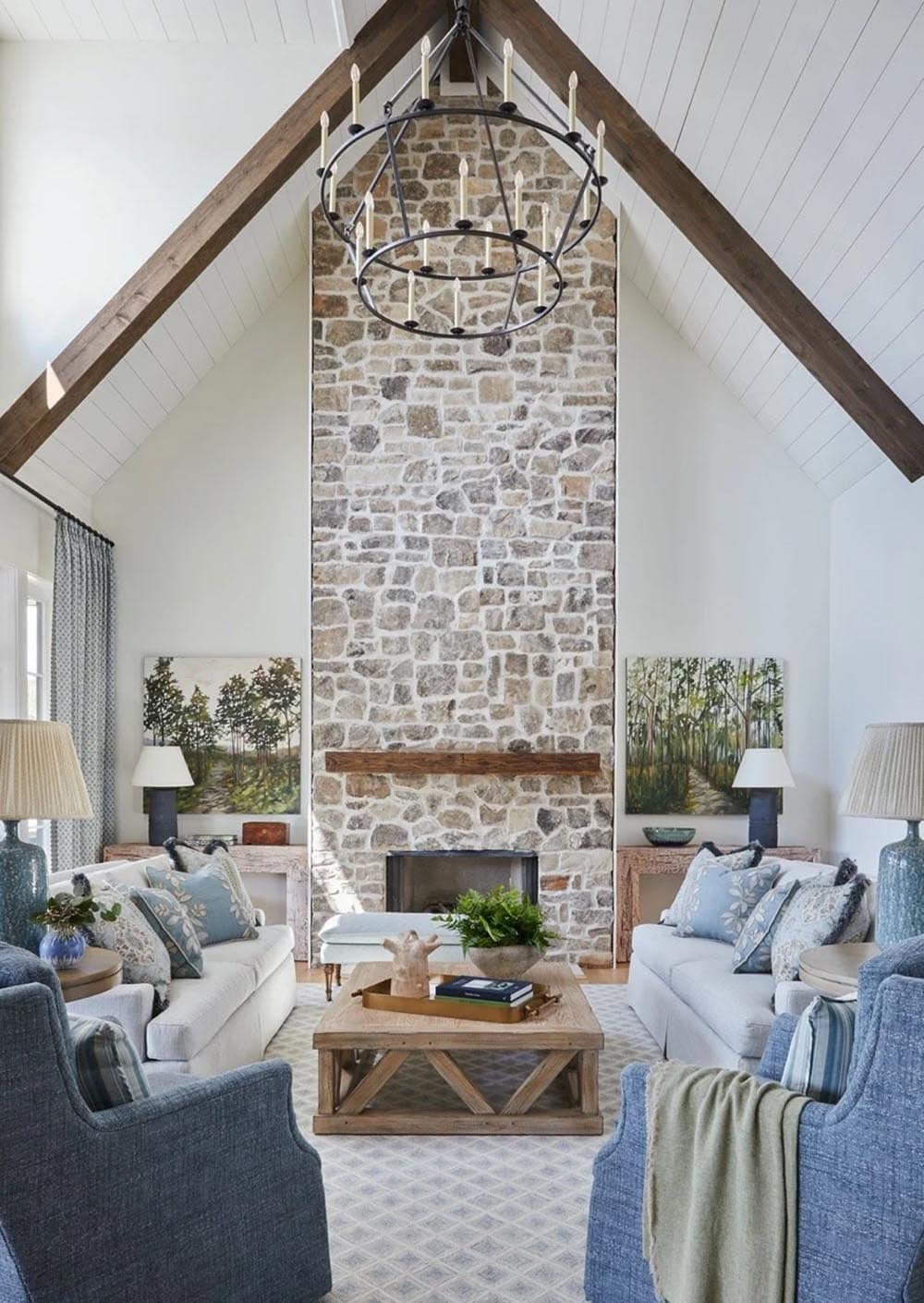 Soaring cathedral ceiling with crossed wood beams, floor-to-ceiling stone fireplace, iron chandelier, and symmetrical seating in blue and white