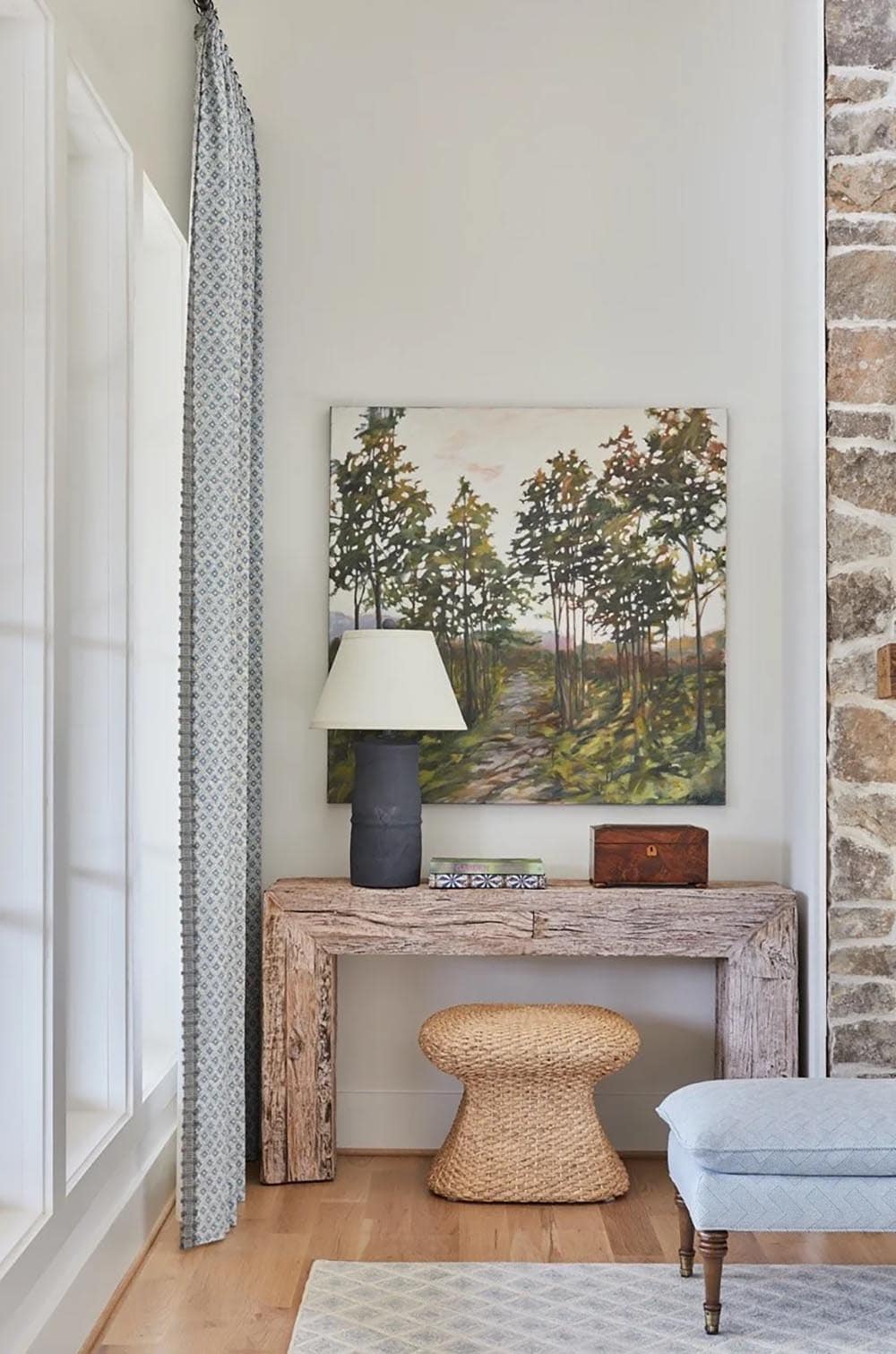 Rustic reclaimed wood console table with landscape painting, woven stool, and patterned curtain beside stone fireplace wall
