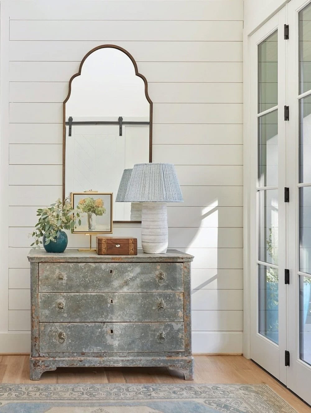 Weathered gray chest of drawers with arched mirror, ceramic lamp, and teal vase against shiplap wall