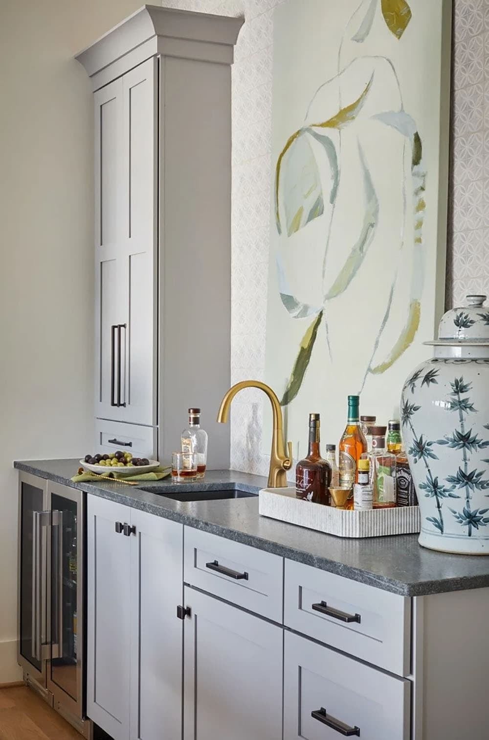 Gray wet bar with dark stone countertop, gold faucet, wine cooler, and large floral painting backdrop