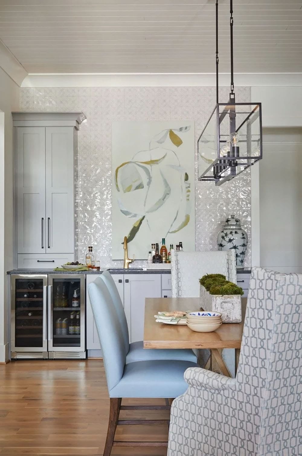 Dining nook with light blue chairs, wooden table, bar area with reflective tile backsplash, and cage pendant light
