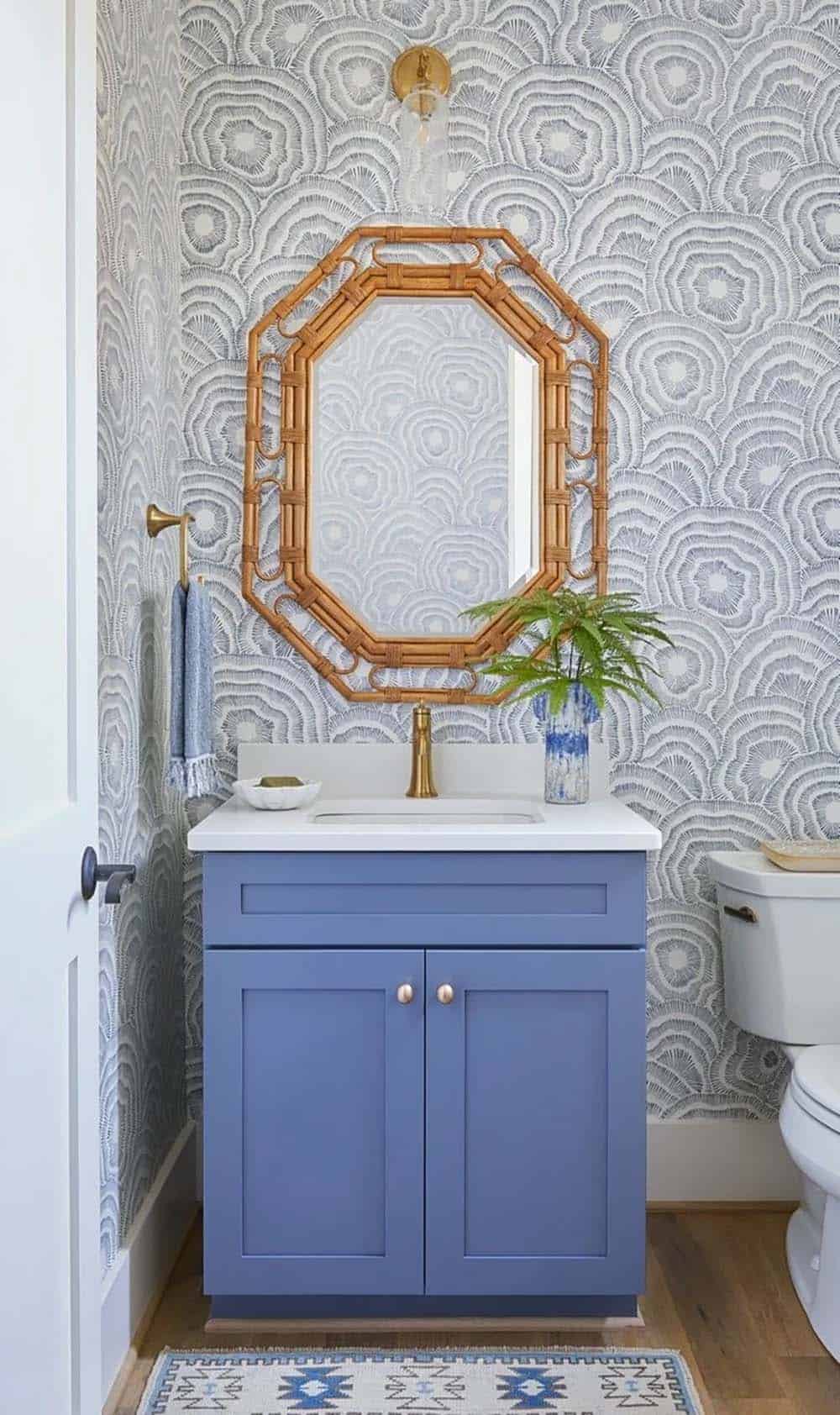 Powder room with bold blue-gray floral wallpaper, cobalt blue vanity, rattan mirror, and gold fixtures
