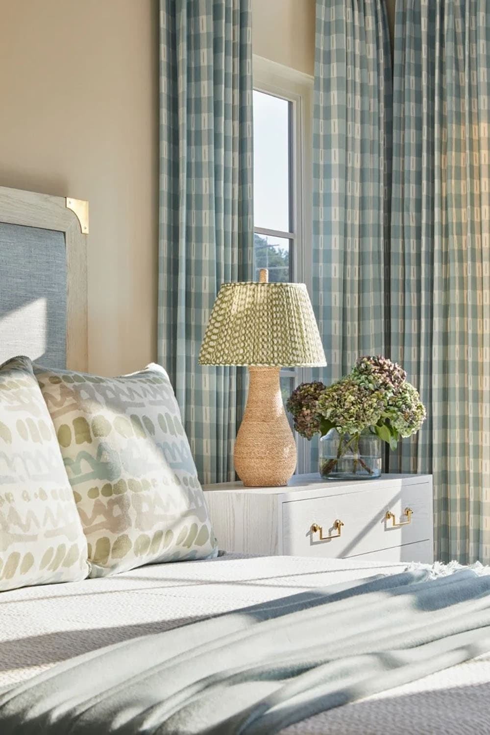 Bedroom detail with woven lamp, plaid curtains, patterned pillows, and dried hydrangeas on nightstand