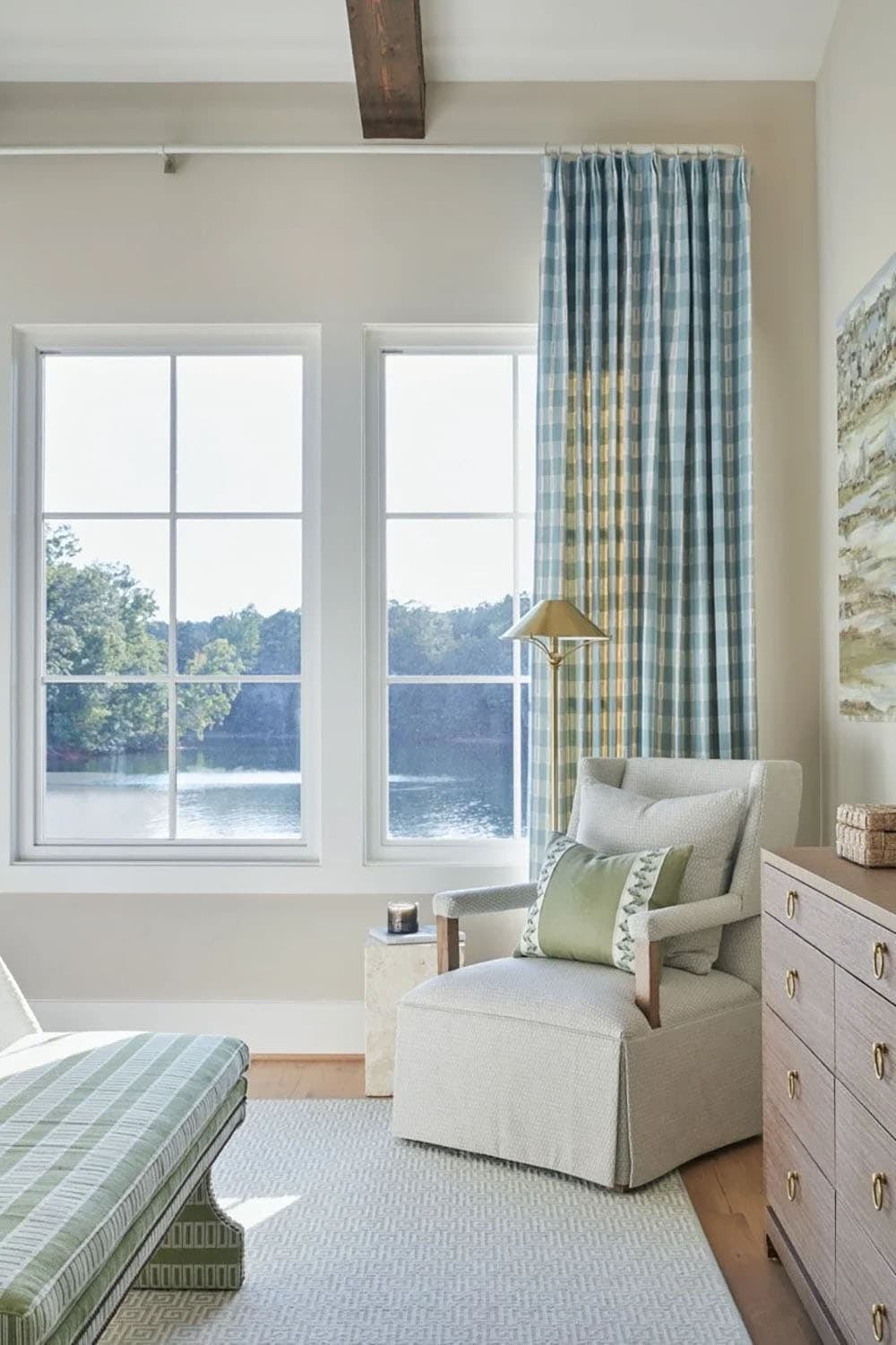 Bedroom sitting area with cream slipper chair, blue plaid curtains framing lake view, and green striped chaise