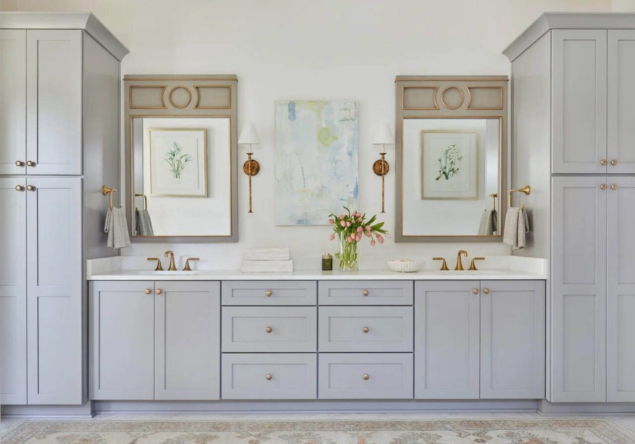 Primary bathroom double vanity with gray cabinetry, gold hardware, decorative mirrors with gold frames, and botanical prints