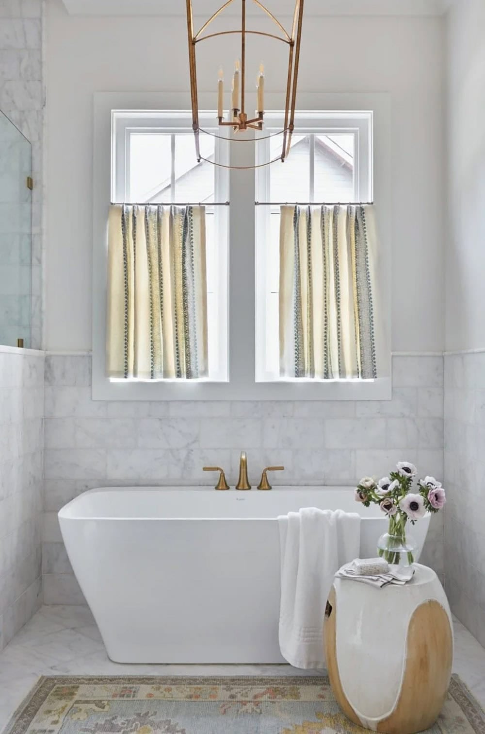 White freestanding soaking tub with gold faucet, marble tile surround, café curtains, gold lantern chandelier, and ceramic garden stool side table