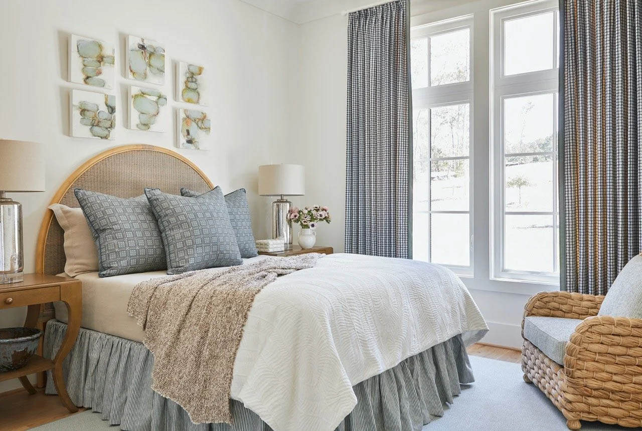 Guest bedroom with arched rattan headboard, blue geometric pillows, plaid curtains, gallery wall of abstract stone paintings, and woven lounge chair