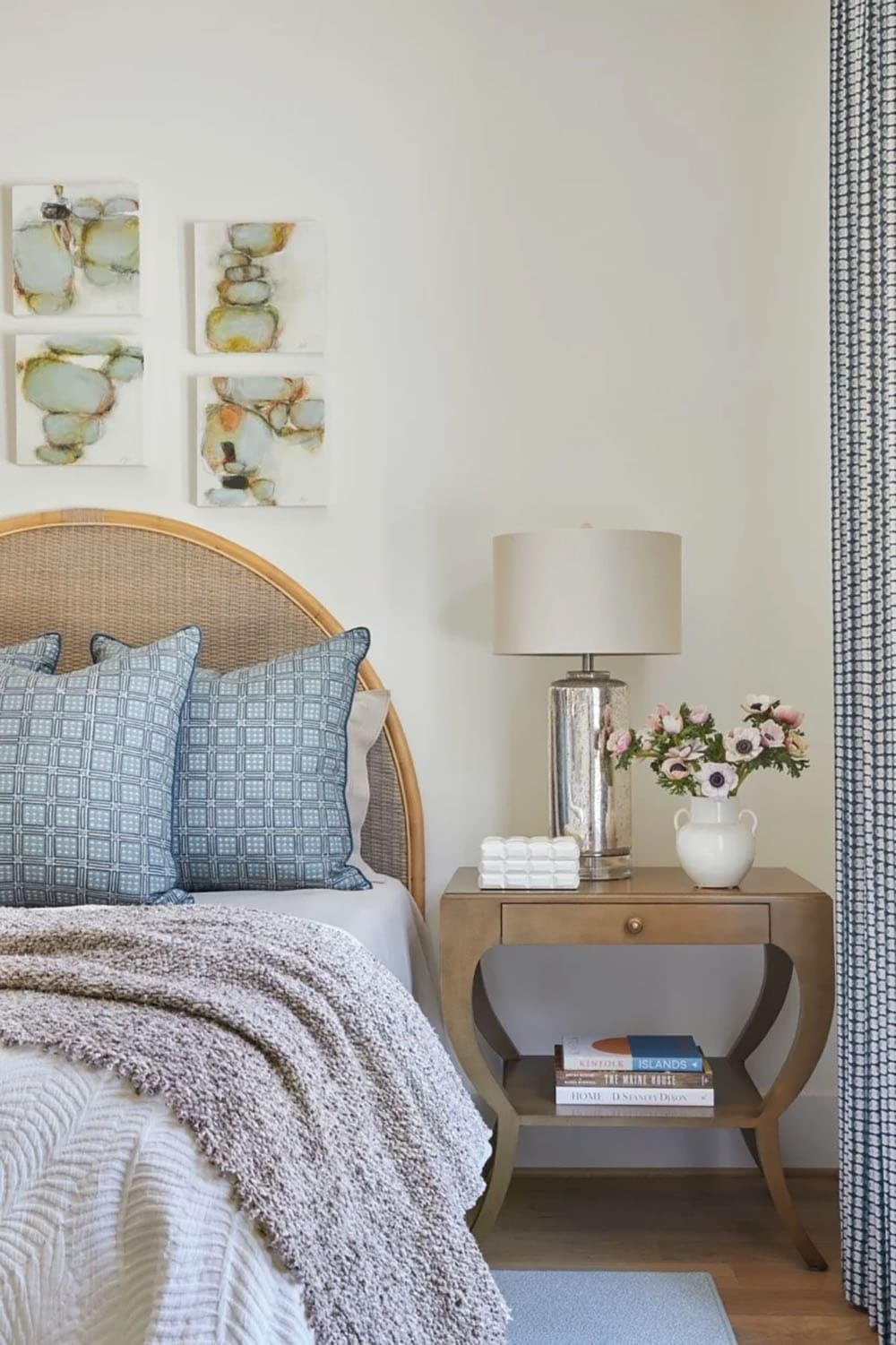 Bedroom nightstand detail with curved wood table, mercury glass lamp, white ceramic vase of anemones, and abstract stone art above arched rattan headboard