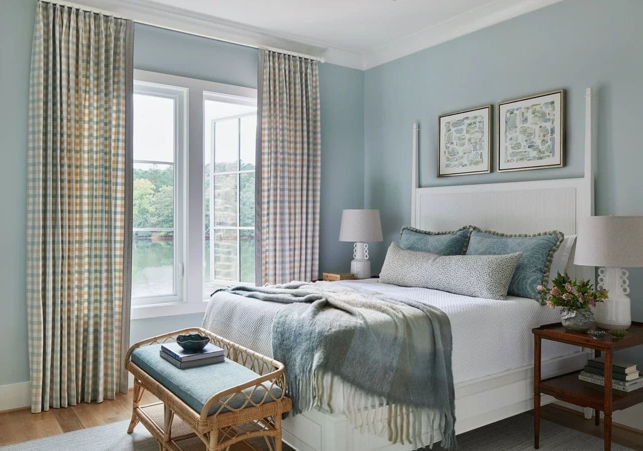 Light blue guest bedroom with white beadboard poster bed, plaid curtains, rattan bench, stacked abstract map prints, and lake views