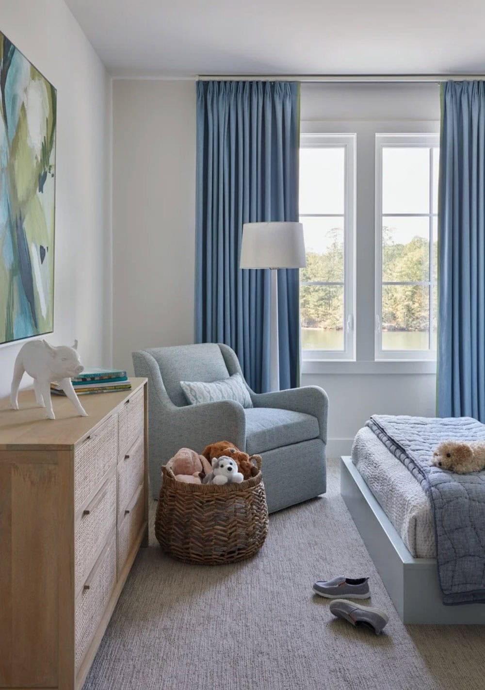 Children's bedroom with blue swivel chair, light wood dresser, basket of stuffed animals, blue curtains, and abstract green painting