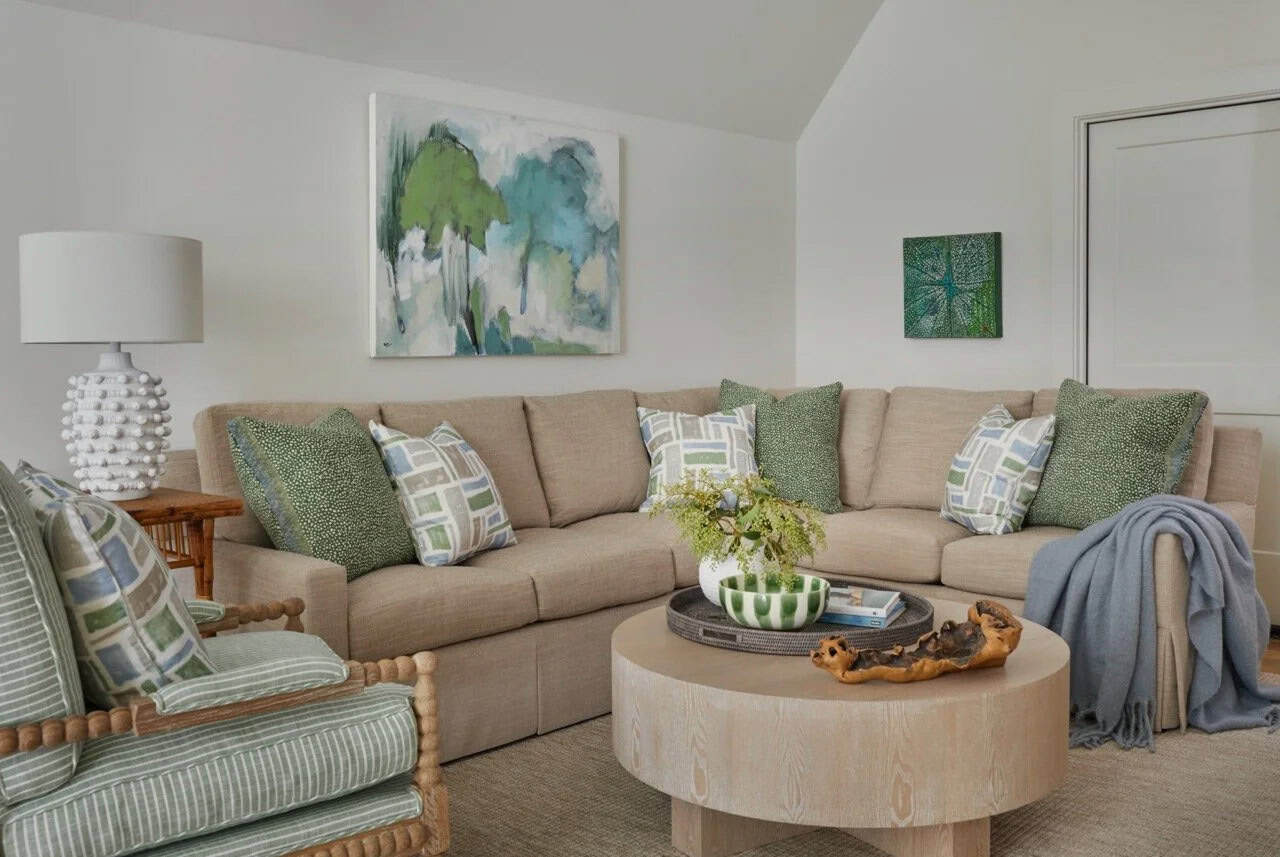 Casual family room with linen sectional, green and blue patterned pillows, round wood coffee table, spool chair, and abstract green tree painting