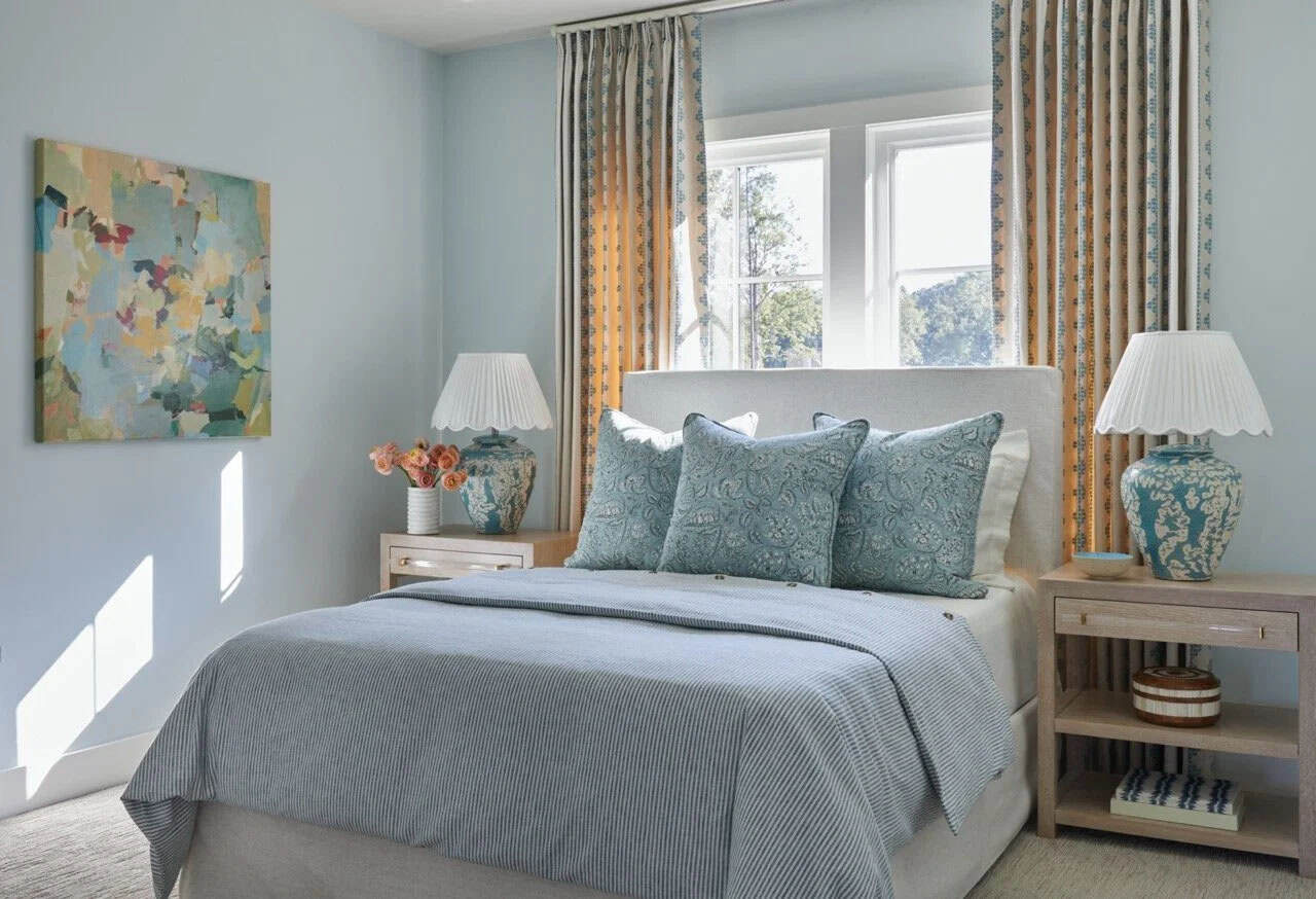 Light blue guest bedroom with blue striped bedding, paisley pillows, patterned curtains, matching ceramic lamps with scalloped shades, and colorful abstract painting