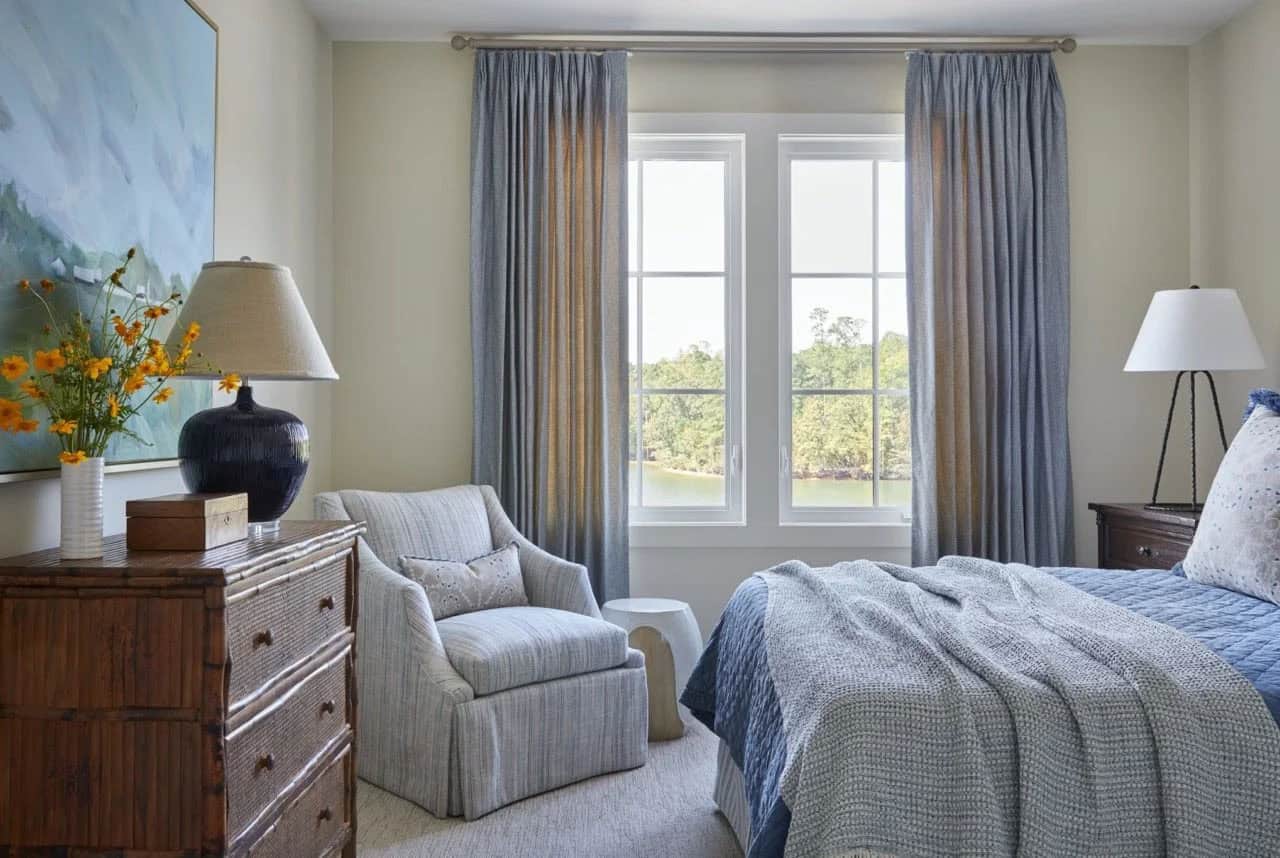 Guest bedroom with blue quilted bedding, gray armchair, bamboo dresser, lake views through blue-gray curtains, and landscape painting