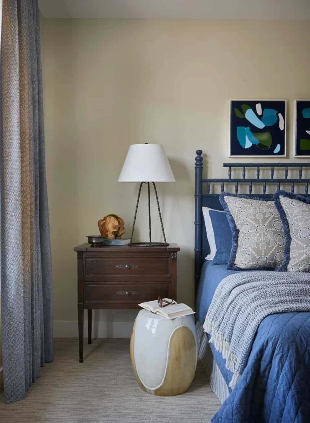 Bedroom nightstand vignette with navy spindle bed, dark wood chest, tripod lamp, ceramic garden stool, and bold blue abstract art