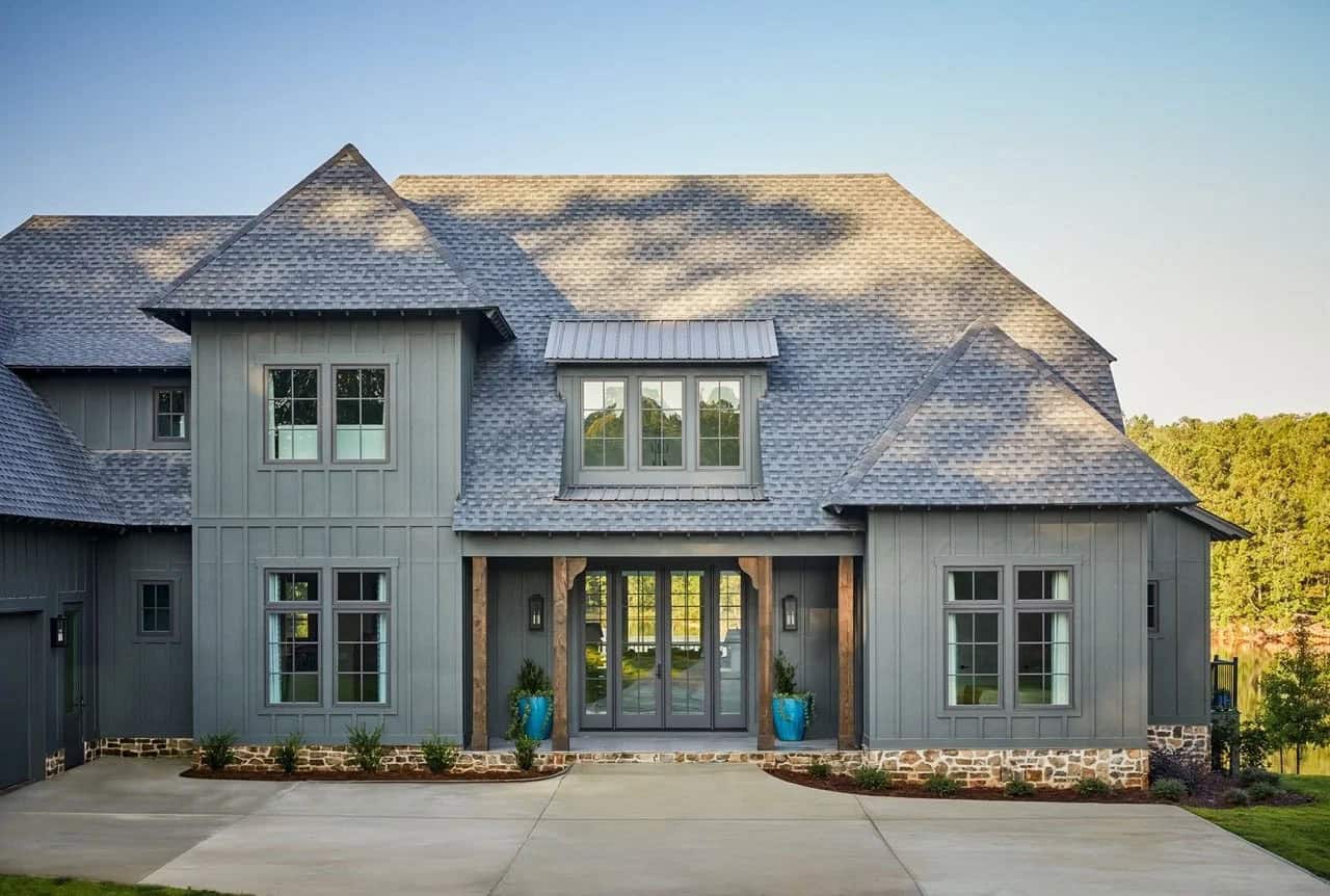Contemporary lake house exterior with gray board-and-batten siding, stone foundation, steep shingle roof, and rustic wood entry columns at dusk
