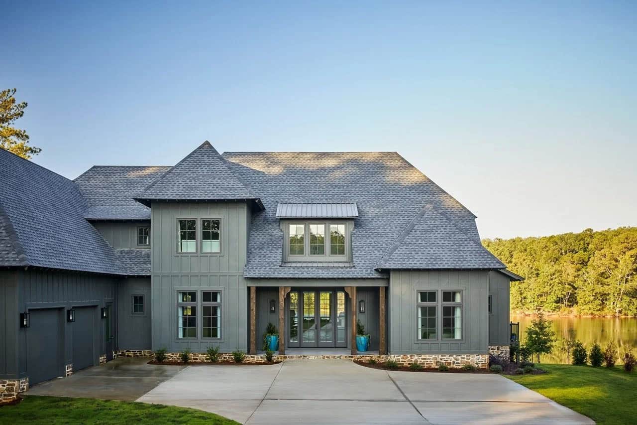Wide exterior view of gray contemporary lake house with stone base, steep multi-gabled roof, wood entry columns, and lake visible in background