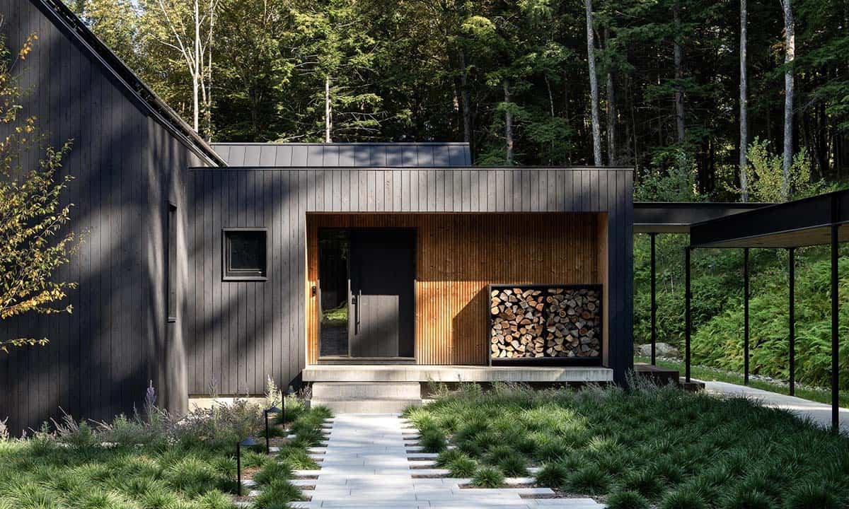 Dark wood exterior of a modern Scandinavian-inspired home in Vermont with a covered entryway, stacked firewood, and lush forest backdrop