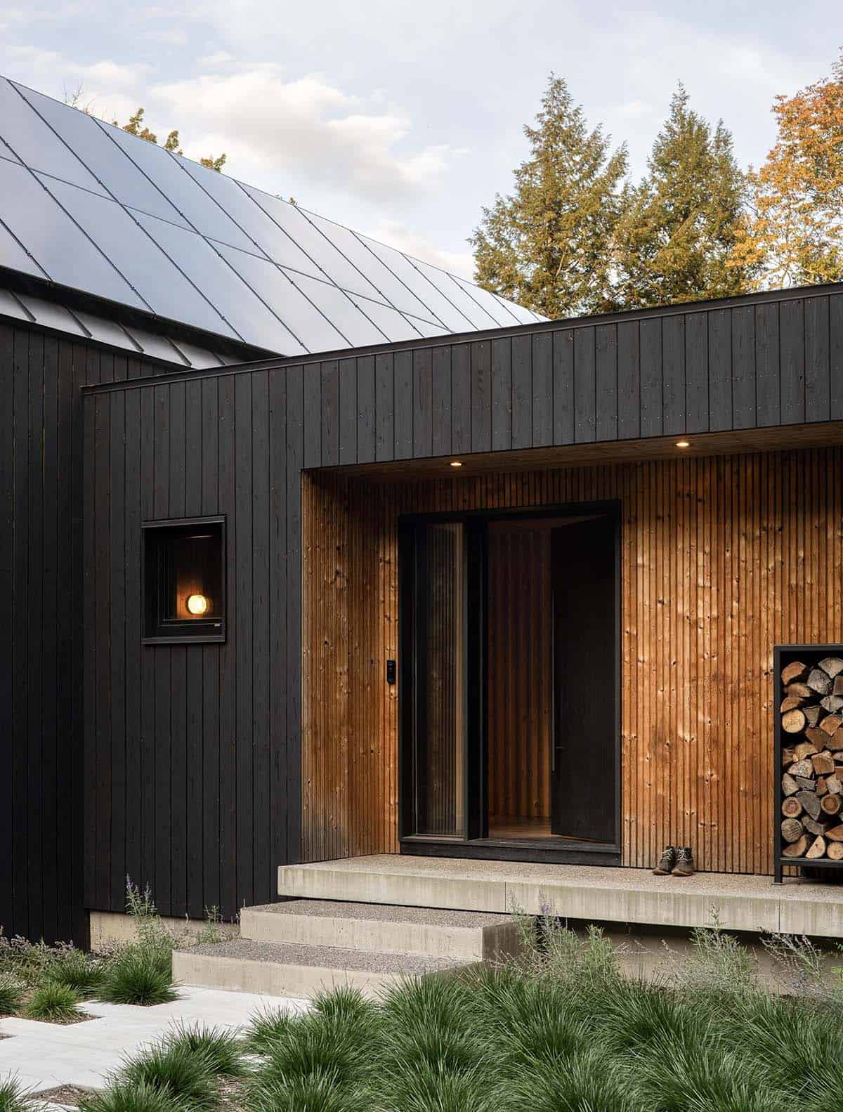 Close-up of the home's dark wood siding, warm cedar entry alcove, solar panel roof, and concrete steps
