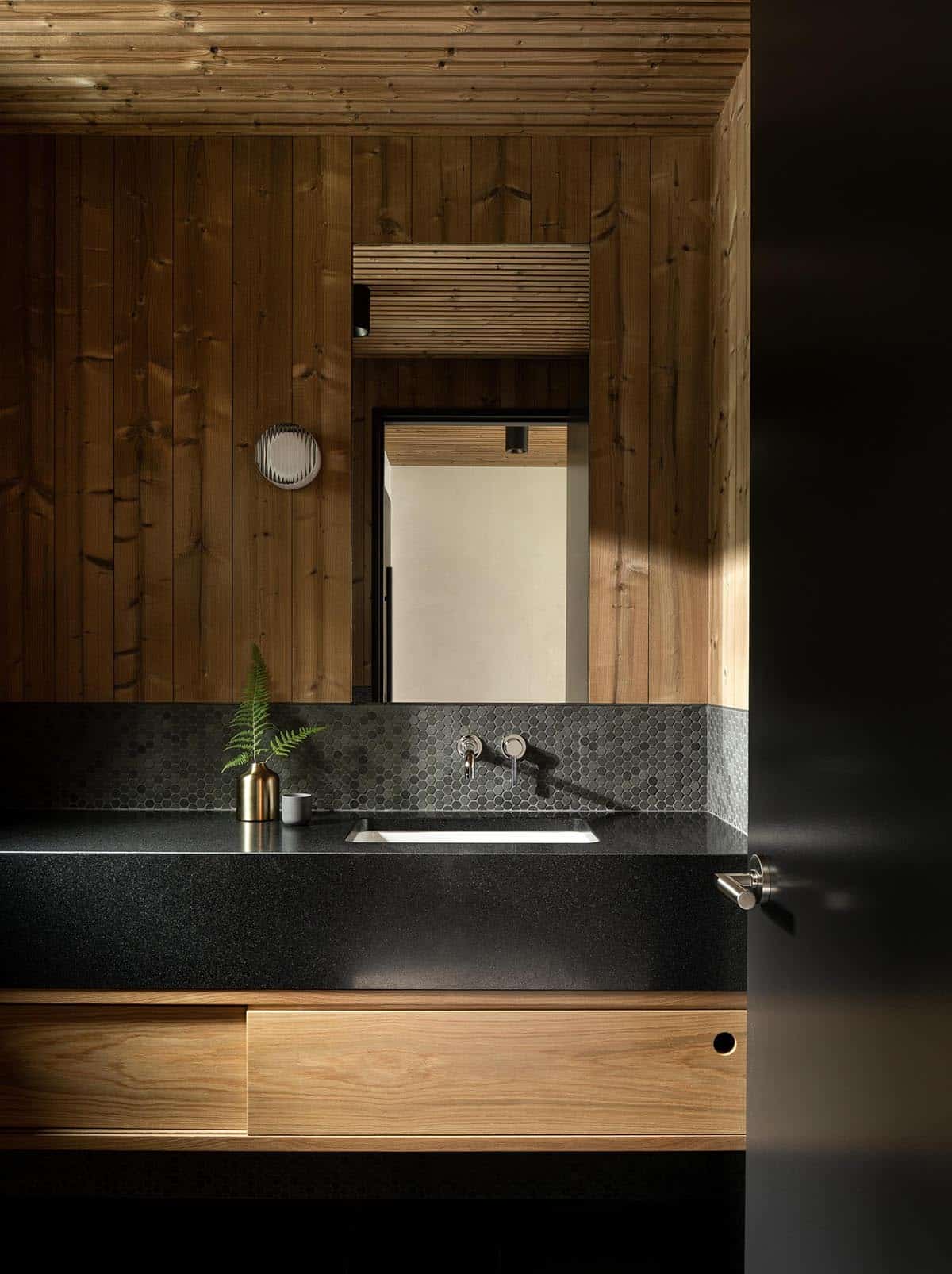 Dark and moody bathroom with knotty pine walls, black granite countertop, hexagonal tile backsplash, and oak vanity