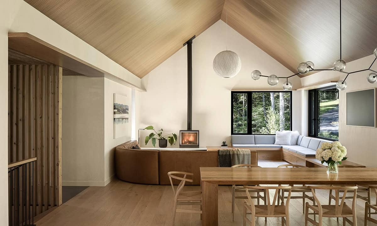 Open-plan living and dining area with cathedral ceiling, wood-burning stove, curved built-in seating, and forest views