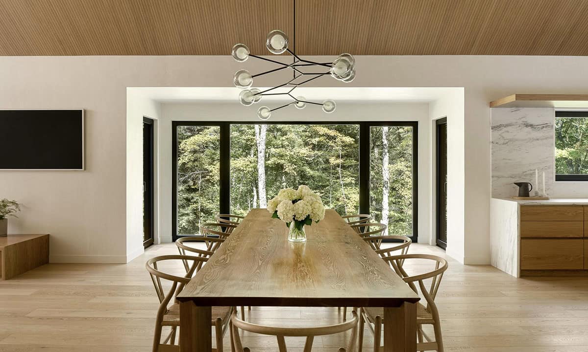 Dining room with long oak table, wishbone chairs, modern branch chandelier, and floor-to-ceiling forest-view windows