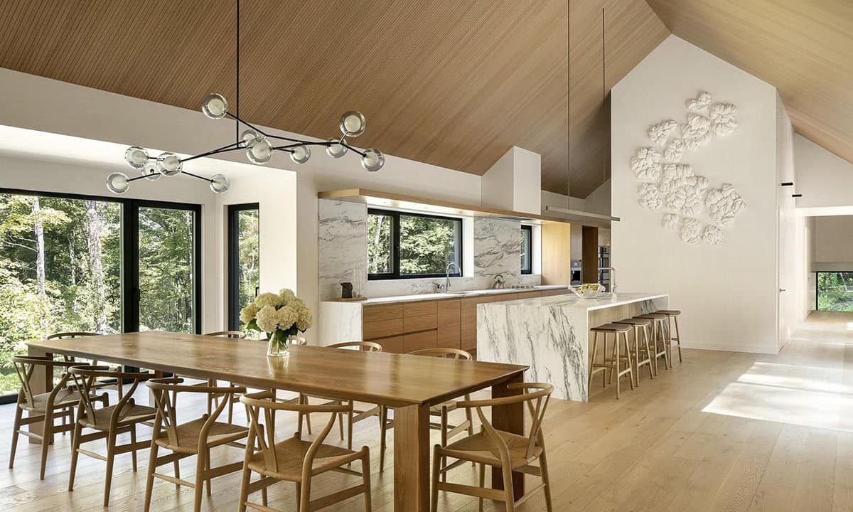 Open kitchen and dining area with vaulted ceiling, marble island, wishbone chairs, and panoramic woodland windows