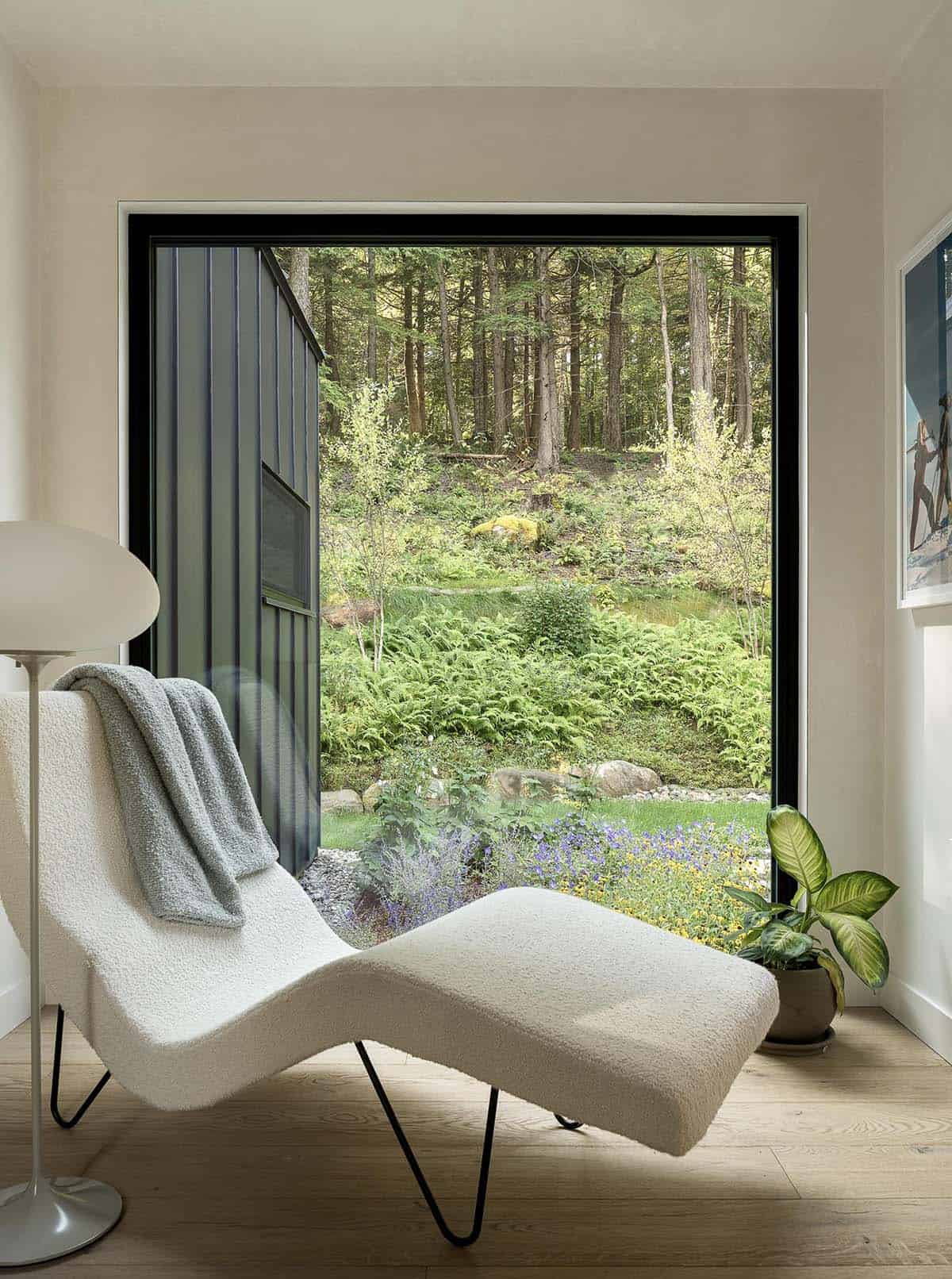 White boucle chaise lounge positioned beside an oversized black-framed picture window overlooking a fern-filled garden
