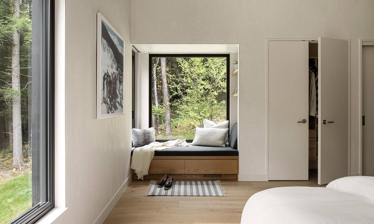 Bedroom corner with an oak window seat and storage drawers framing a large black-framed forest view window