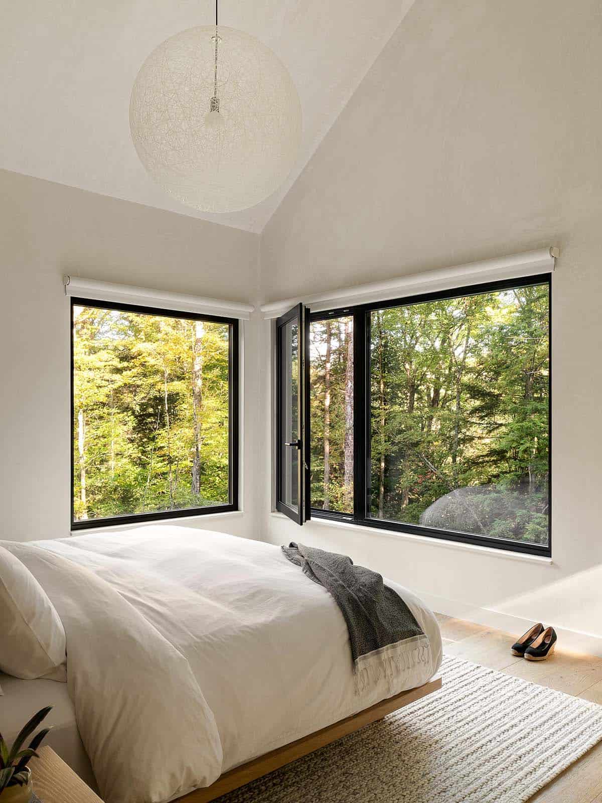 Minimalist primary bedroom with corner black-framed windows opening to forest canopy and a white woven globe pendant