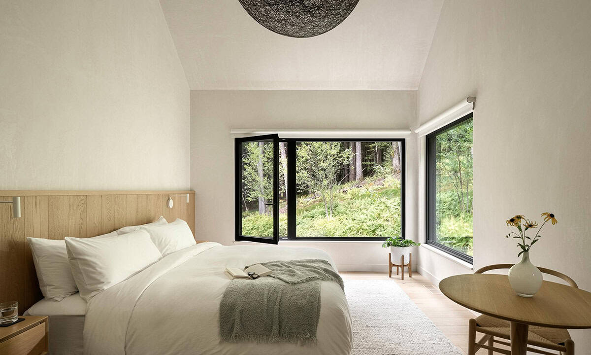 Serene primary bedroom with oak headboard, white bedding, dark woven pendant, and corner windows overlooking greenery