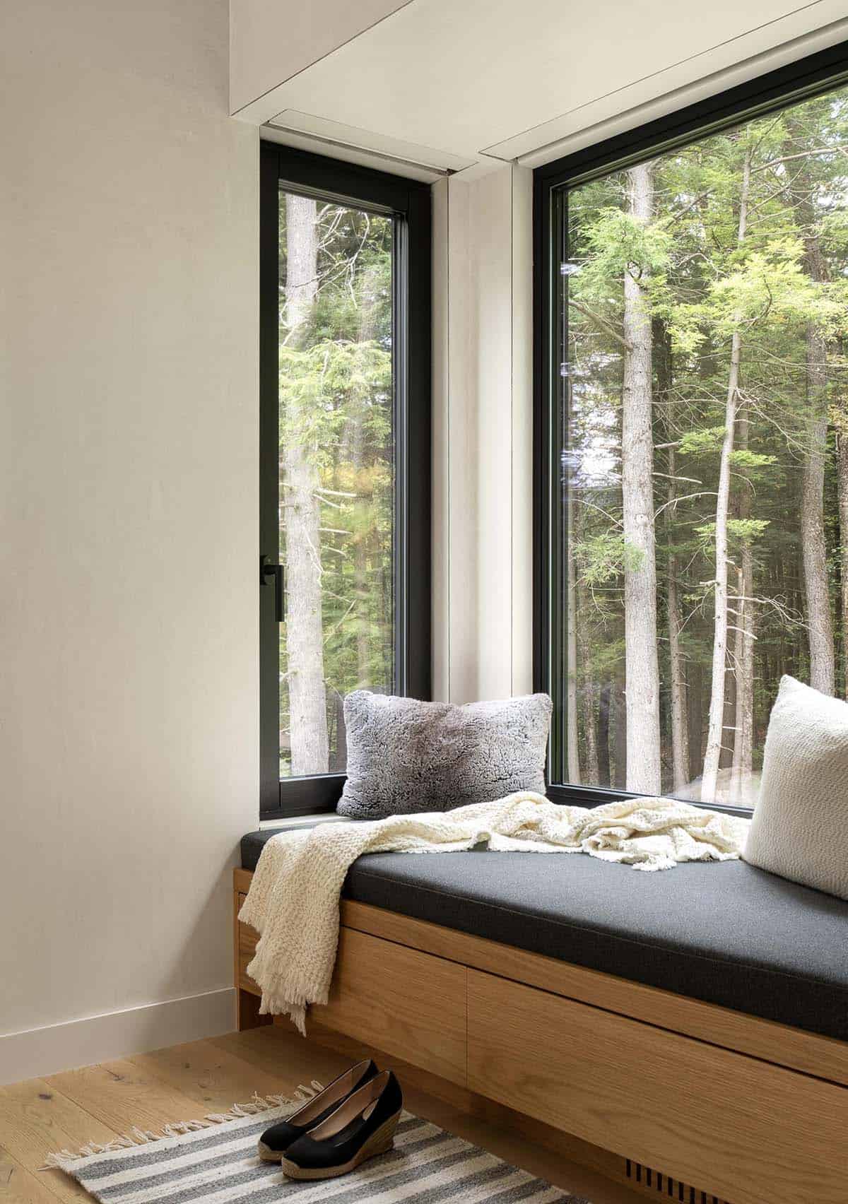 Built-in oak window seat with dark cushion and throw pillows nestled in a corner of floor-to-ceiling forest windows