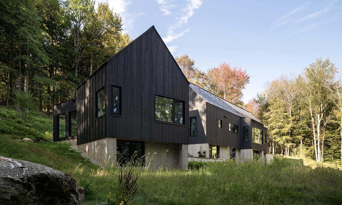 Side view of a dark-clad contemporary Vermont home with gabled roof, concrete foundation, and large black-framed windows set into a wooded hillside