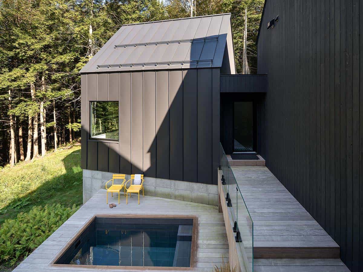 Rooftop plunge pool with wood deck, two yellow chairs, and glass railing beside a dark metal-clad gable structure