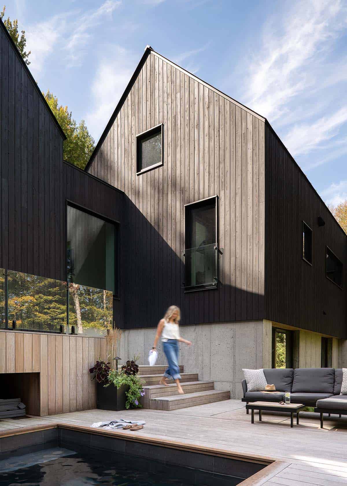Person walking up wood and concrete pool deck steps beside a dark cedar gable home with outdoor lounge seating