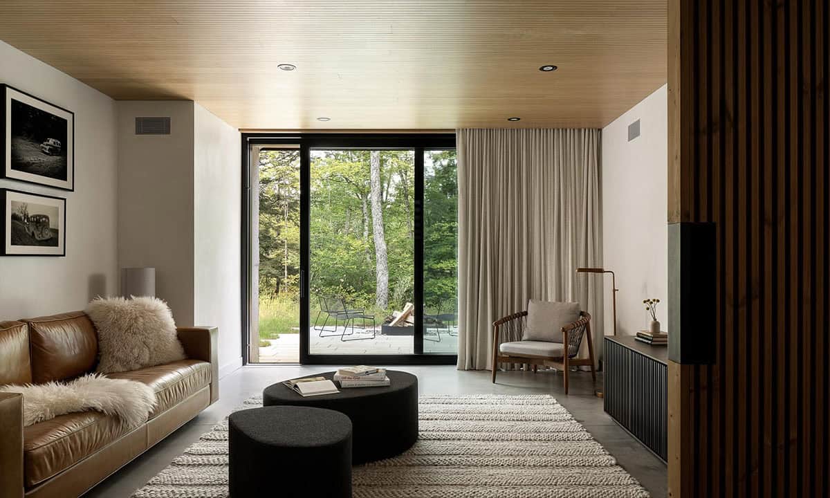 Lower-level living room with wood-paneled ceiling, leather sofa, round black ottomans, linen curtains, and sliding glass doors to a garden