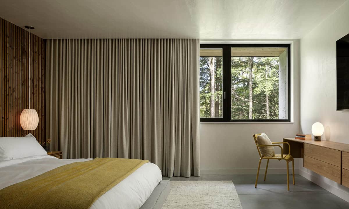 Bedroom with floor-length linen curtains, mustard yellow throw, slatted wood accent wall, globe pendant light, and forest window view
