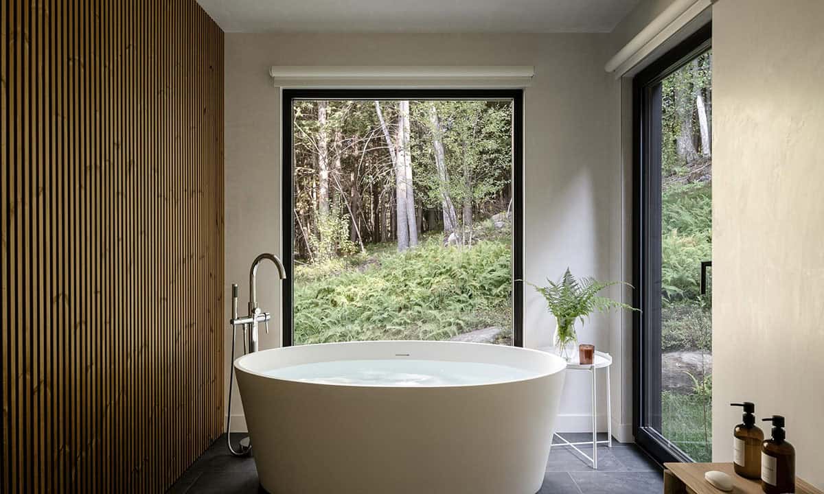 Freestanding white soaking tub centered before a large black-framed picture window overlooking a lush fern garden