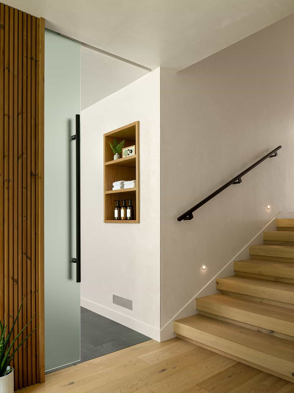 Frosted glass sliding door beside a built-in oak niche with towels and toiletries, adjacent to a light wood open staircase