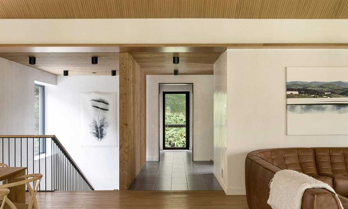 Upper hallway with wood-clad ceiling portal framing a forest view window, feather artwork, and glimpse of a leather sofa