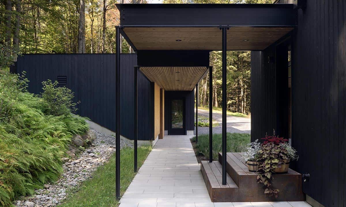 Covered outdoor walkway with black steel posts, cedar ceiling, and stone path connecting building wings through a wooded landscape