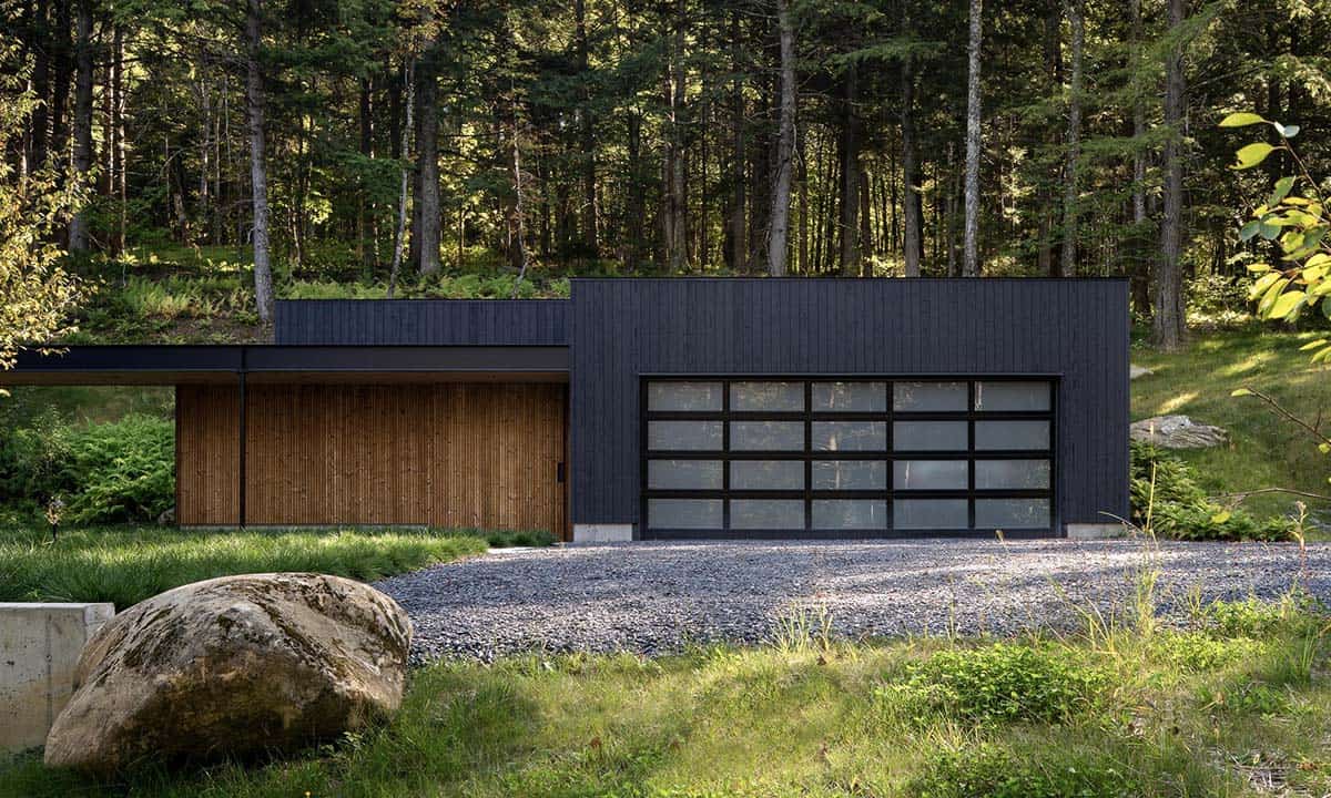 Dark wood garage structure with a full-panel glass garage door and warm cedar-clad storage section set against a forest backdrop
