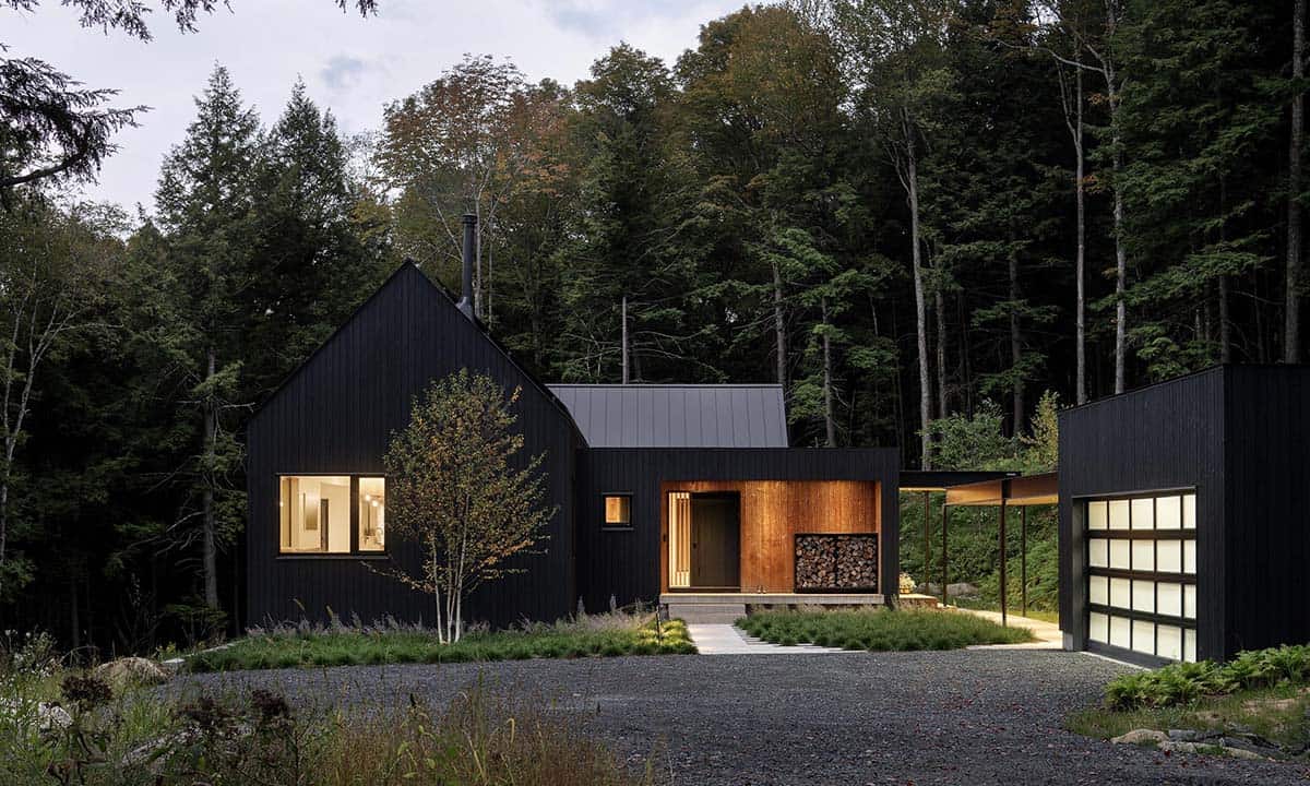 Dusk exterior of the Vermont home with glowing entry alcove, illuminated garage door, and dark wood siding amid autumn trees