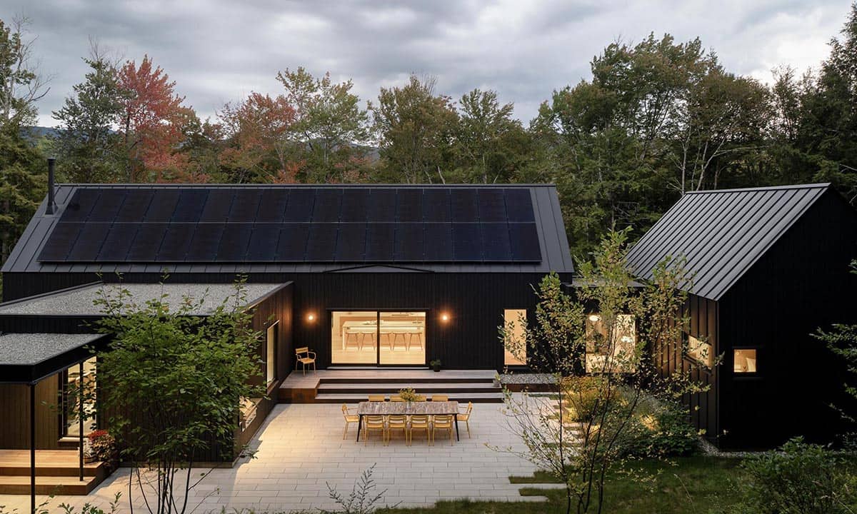 Aerial dusk view of the home's courtyard with solar-paneled roof, yellow outdoor dining set, and warm kitchen light spilling through glass doors
