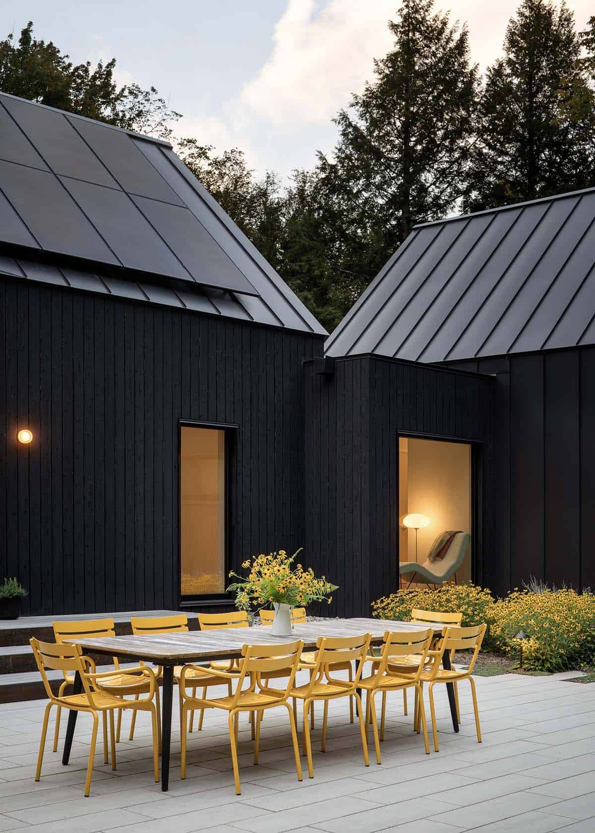 Twilight courtyard view of two dark gabled structures with yellow dining chairs, warm window glow, and blooming yellow garden plantings