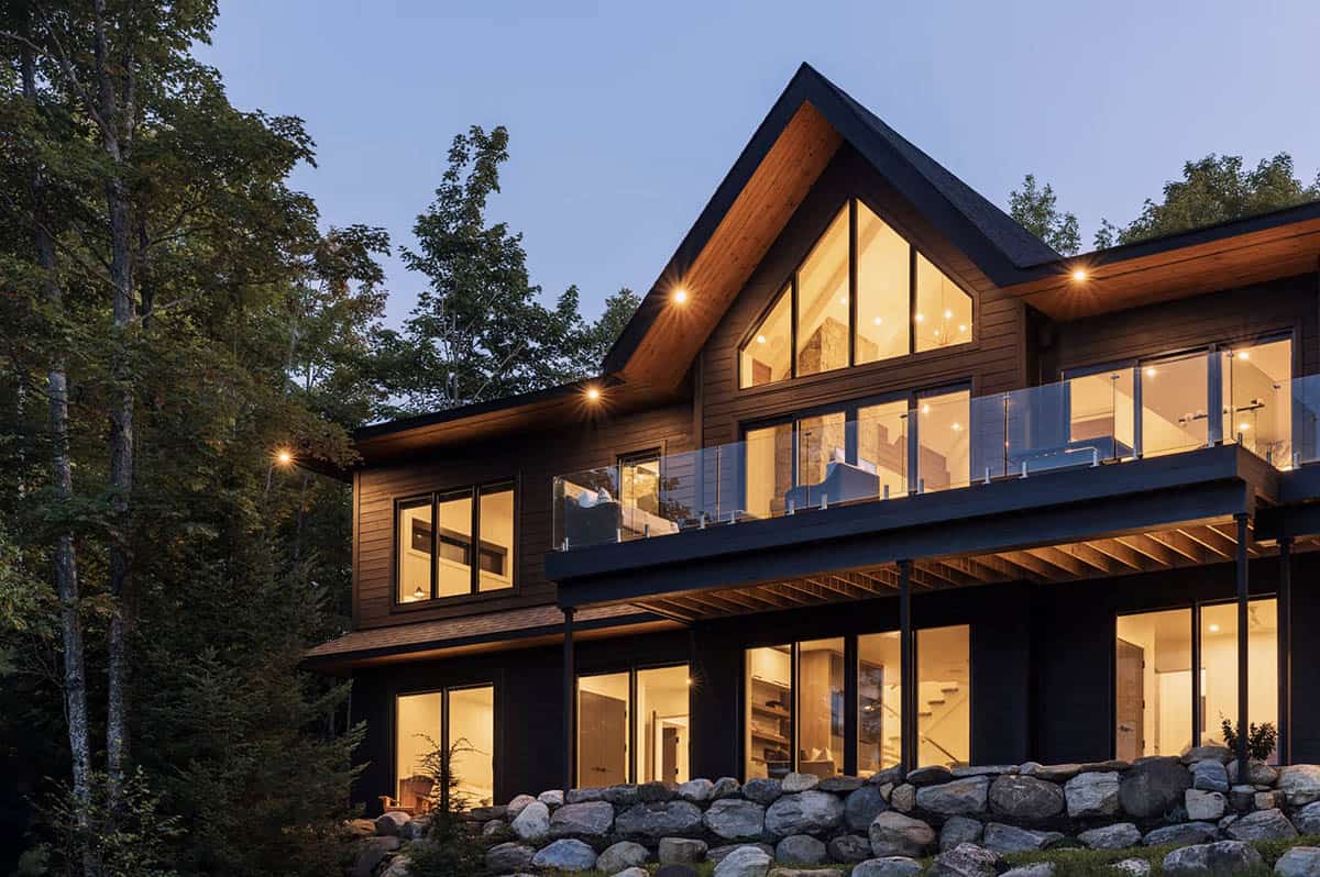 Modern cottage exterior at dusk with warm glowing windows, dark wood siding, and stone foundation